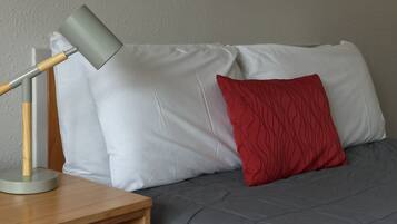 A bed with a grey duvet and a red pillow, a wooden bedside table with a lamp, and a plain wall in the background.