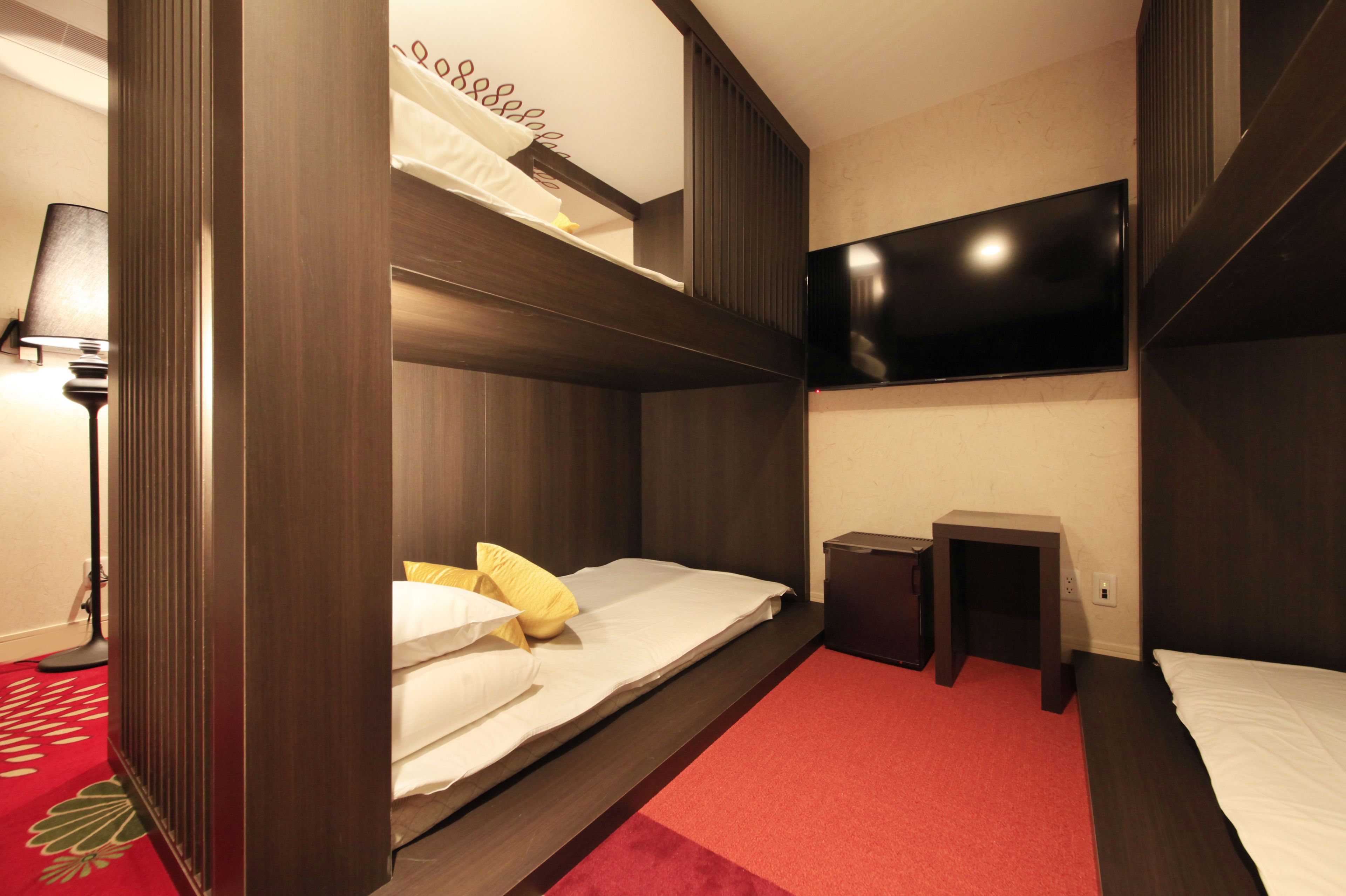 Une chambre avec un lit superposé, un téléviseur fixé au mur, une petite table et une moquette rouge.