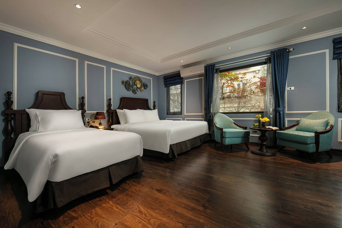 Habitación de hotel con dos camas, una zona de estar con una silla y una mesa, y una ventana con cortinas.