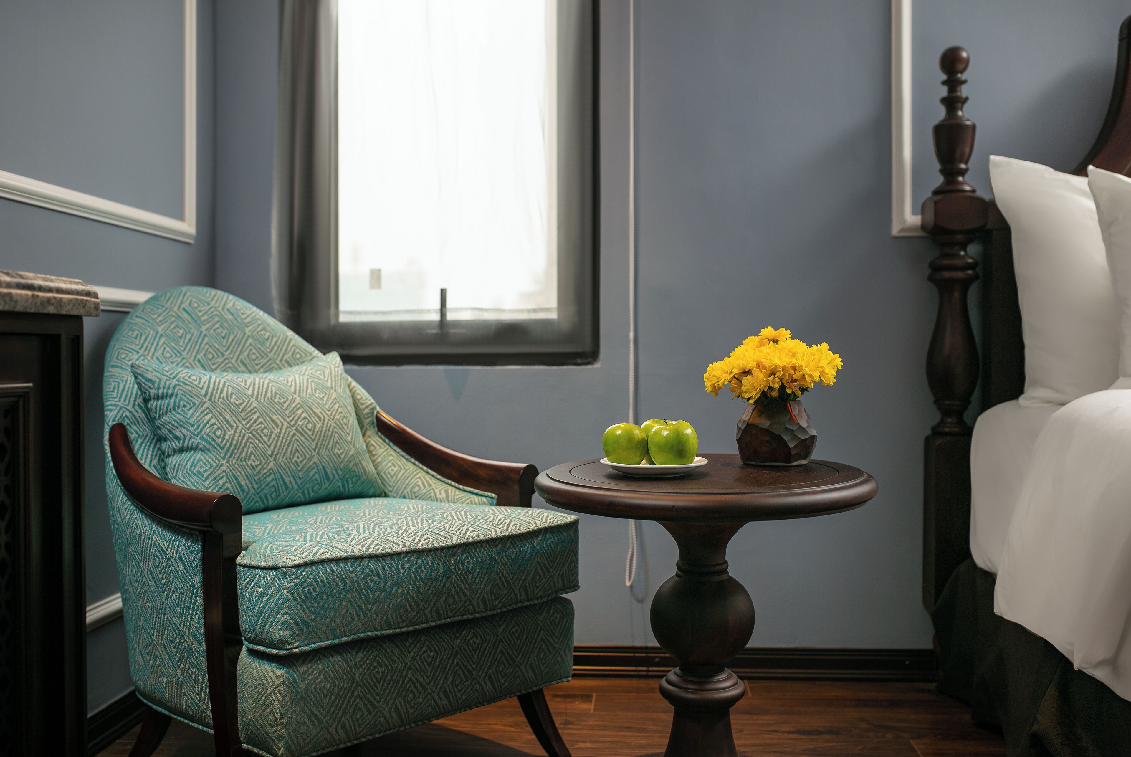 A room with a patterned armchair, a small round table with apples and flowers, and a bed with white linens.