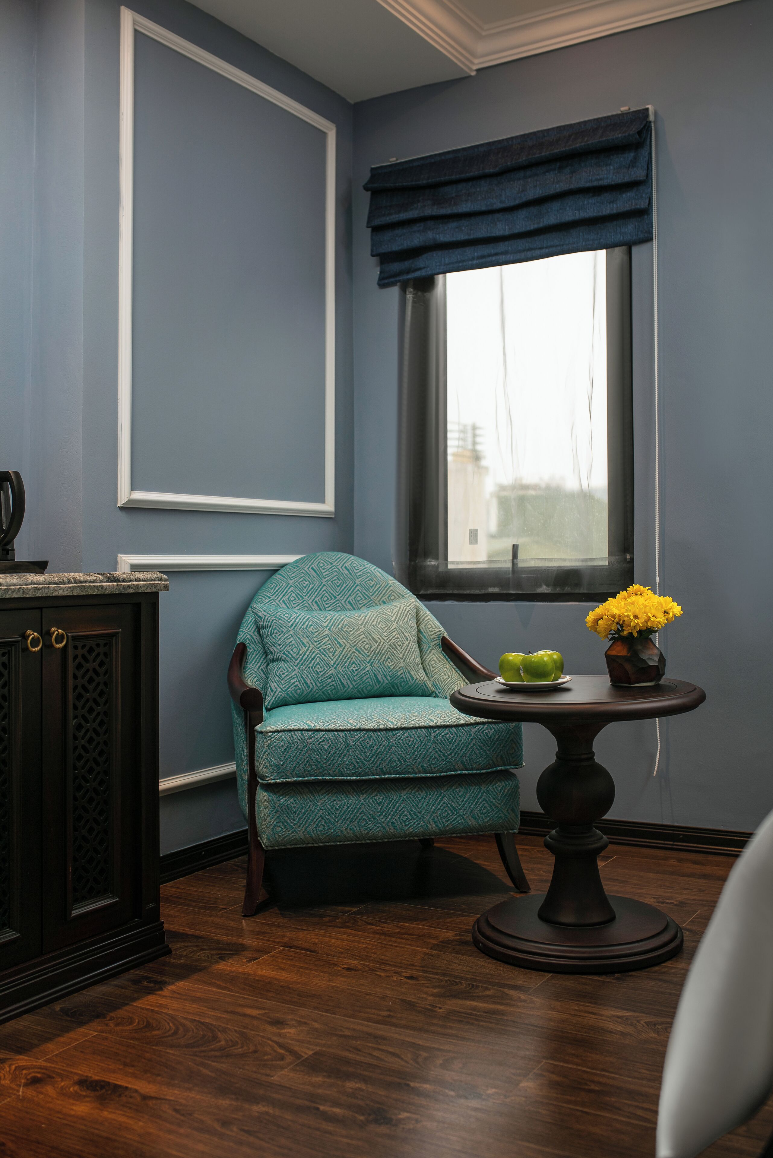 A room with a teal patterned armchair, a small round table with a vase of yellow flowers, and a window with dark blue curtains.
