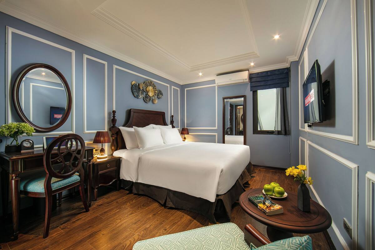Habitación de hotel con una cama grande, un escritorio con una silla, un espejo redondo, un televisor y una mesita con un florero y manzanas.