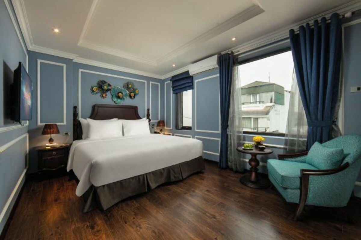 Een hotelkamer met een groot bed, een stoel, een klein tafeltje met bloemen en een uitzicht op gebouwen door het raam.