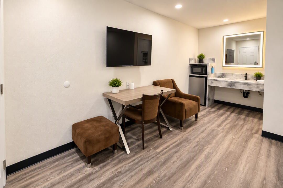 A compact room with a wooden dining table, two chairs, a small sofa, a flat-screen TV mounted on the wall, a microwave, and a sink with a mirror.