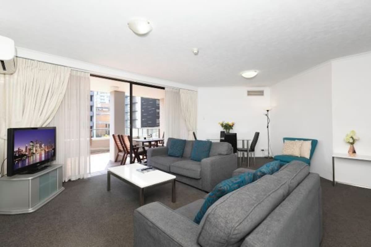 A modern living room with a flat-screen TV, grey sofas, a coffee table, and a dining area with chairs.