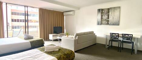 Ein Hotelzimmer mit zwei Betten, einem Schlafsofa, einem Fernseher, einem Schreibtisch mit Stühlen und einem Bild an der Wand.