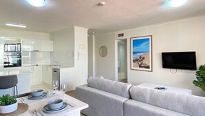 A modern living room with a grey sofa, a dining table set for two, and a kitchen area with white cabinets and appliances.