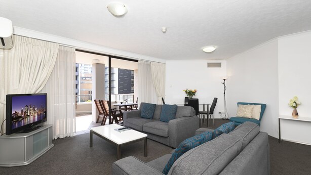 A modern living room with a grey sofa set, a flat-screen TV, a coffee table, and a dining area with chairs and a table.