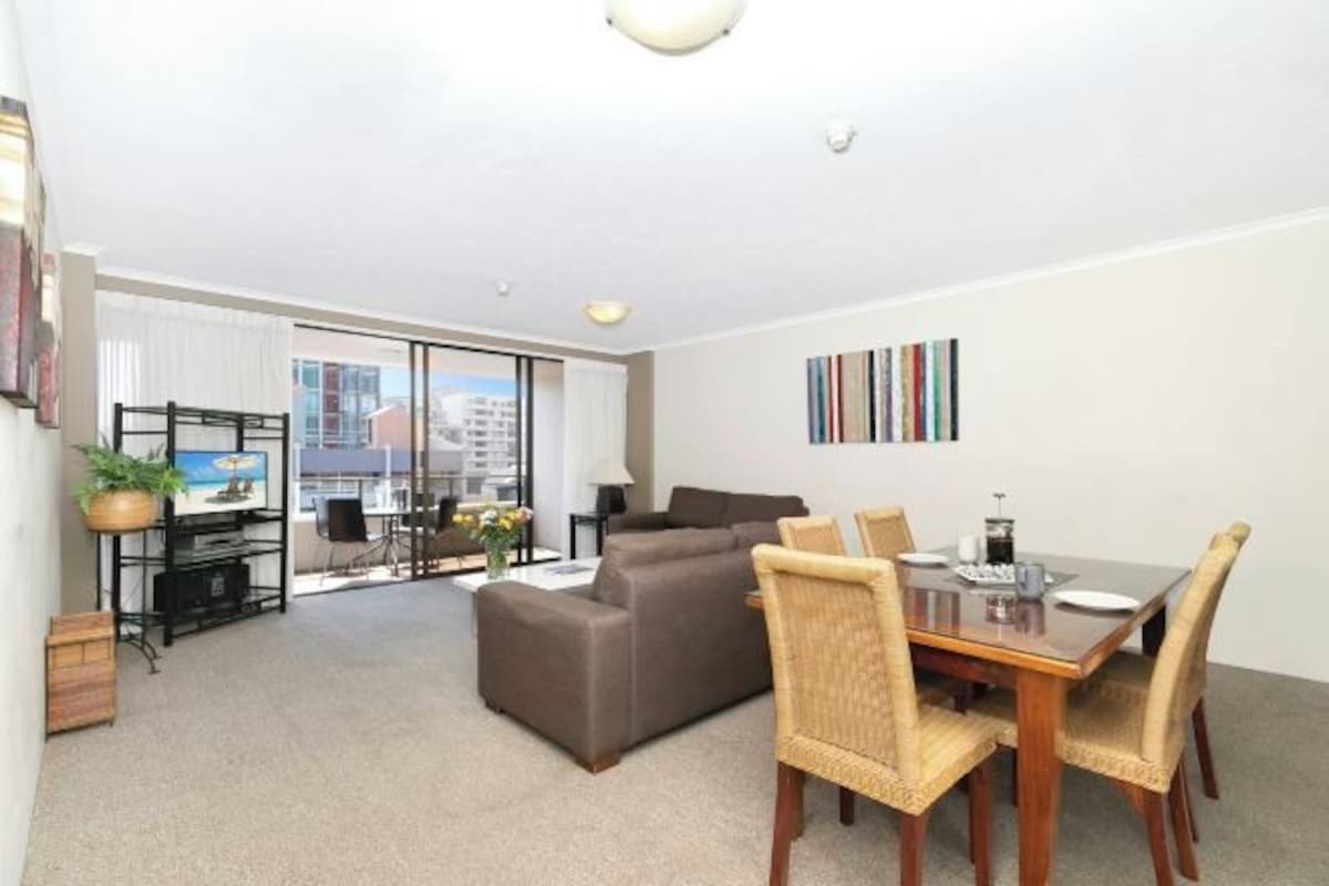 A modern hotel room with a sofa, dining table, chairs, a TV, and a large window with a city view.
