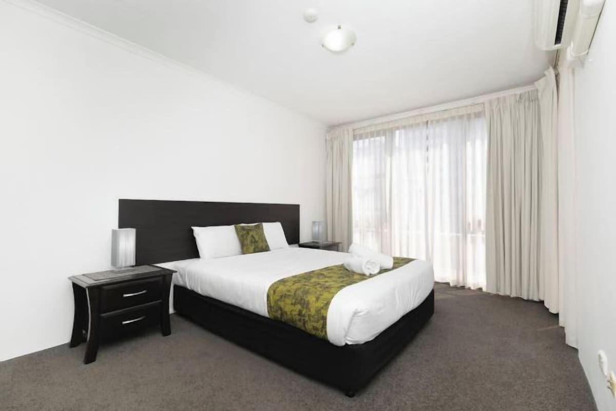 A hotel room with a large bed, a bedside table with drawers, a lamp, and a window with curtains.