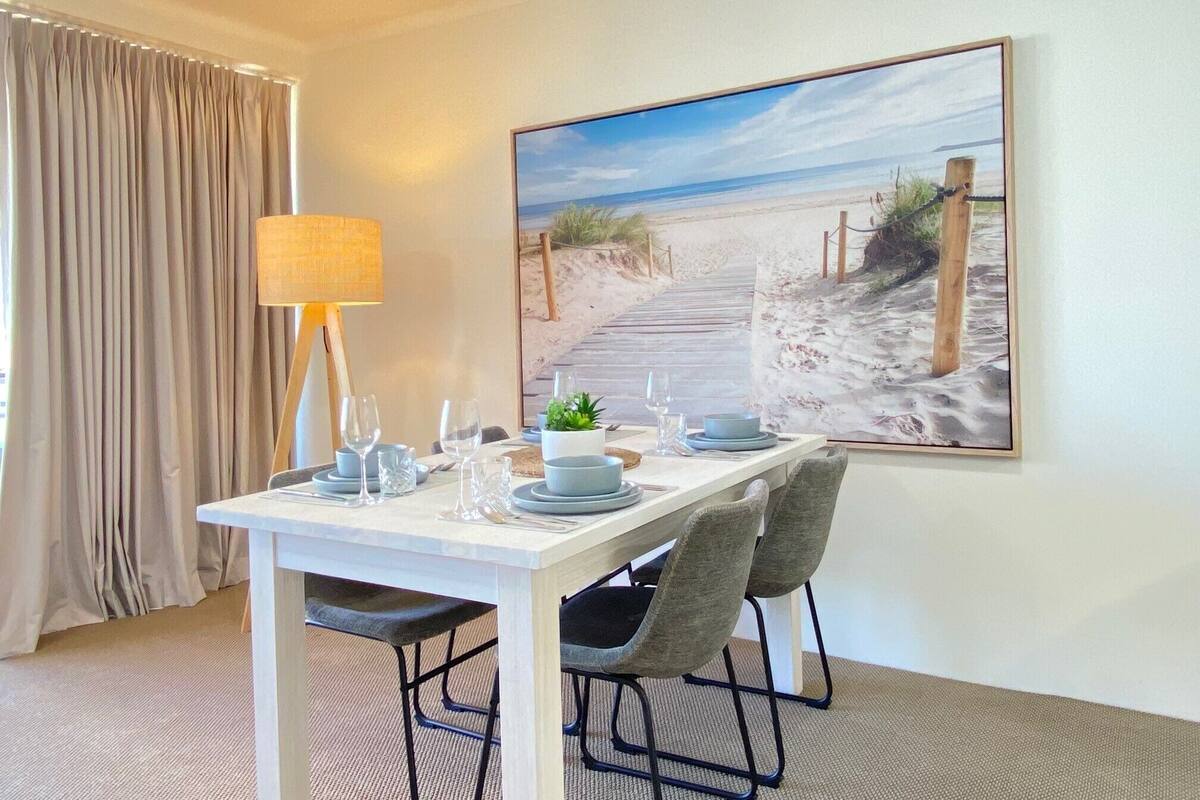 Un comedor con una mesa preparada para cuatro, una lámpara y un cuadro con motivos playeros en la pared.