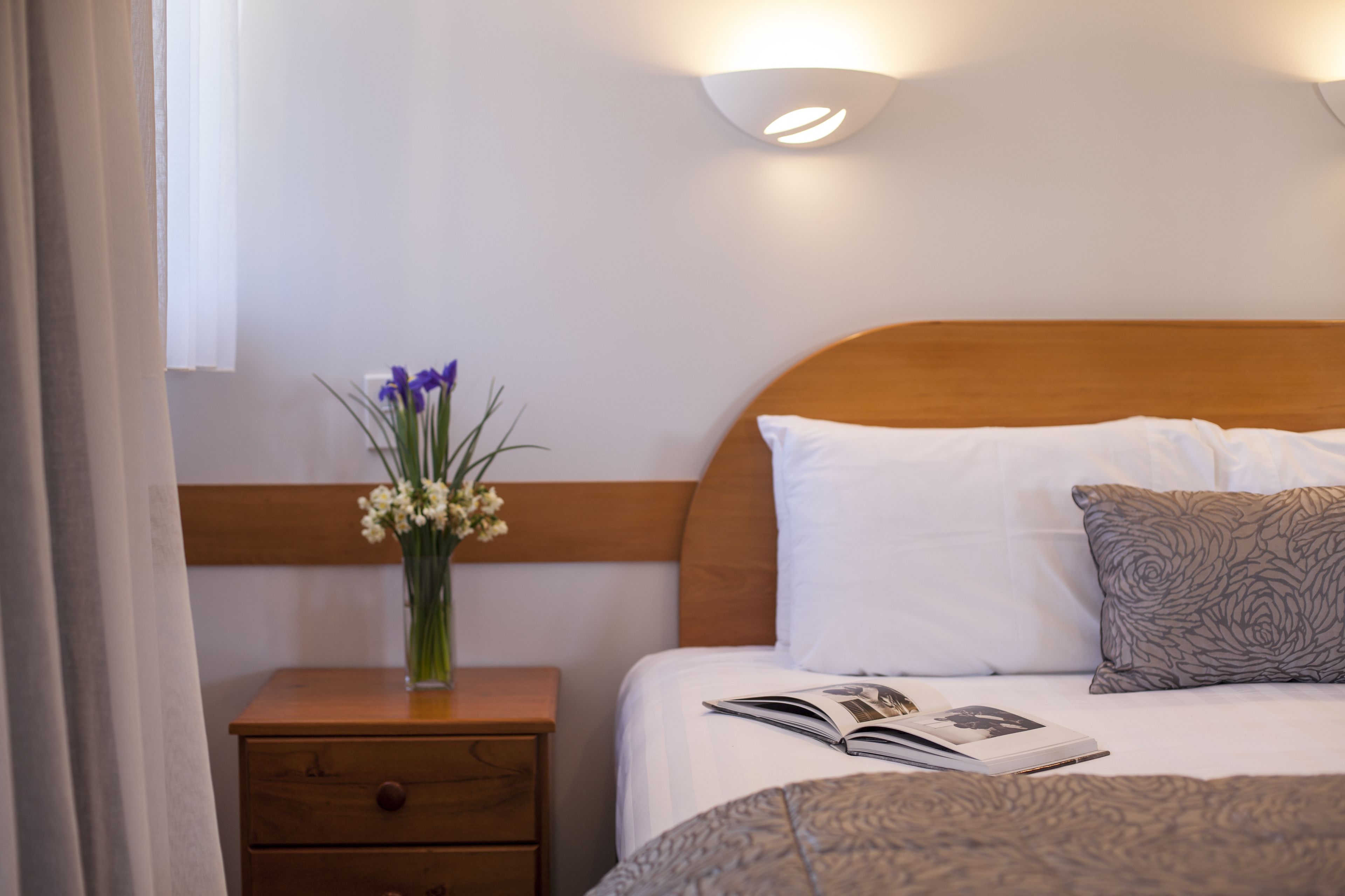 Un lit bien fait, avec une tête de lit en bois, une table de chevet ornée d’un vase de fleurs et un luminaire fixé au mur.