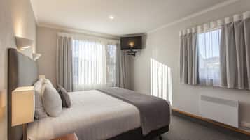 Quarto de hotel moderno com cama, mesa de cabeceira, luminária, TV e janelas com cortinas.