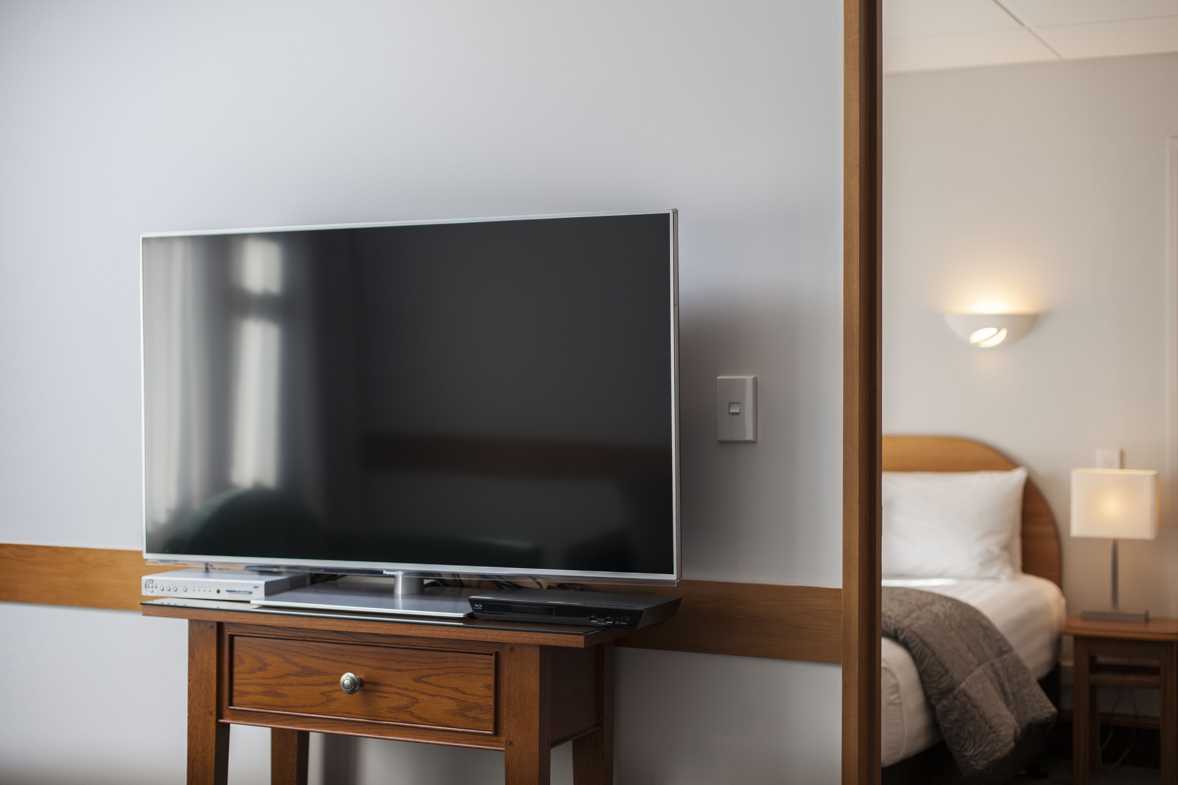 Una camera d'albergo con una TV a schermo piatto su un supporto in legno, un comodino con una lampada e un letto con testiera bianca.