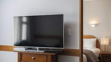 Una camera d'albergo con una TV a schermo piatto su un supporto in legno, un comodino con una lampada e un letto con testiera bianca.