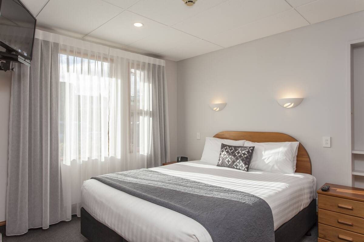 Una habitación de hotel con una cama grande, una mesita de noche de madera, un televisor montado en la pared y dos lámparas fijadas a la pared.