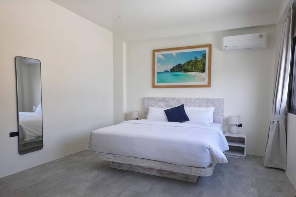 Una habitación de hotel con cama, un cuadro de una playa, un espejo y un equipo de aire acondicionado.