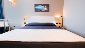 A neatly made bed with a white comforter and a dark blue pillow, a white headboard, and a painting of a sunset over water on the wall. - Hotel Mediterraneo (Villa Cortese)