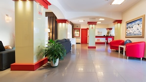A hotel lobby with a reception desk, red and white decor, a grey sofa, and a potted plant. - Hotel Mediterraneo (Villa Cortese)