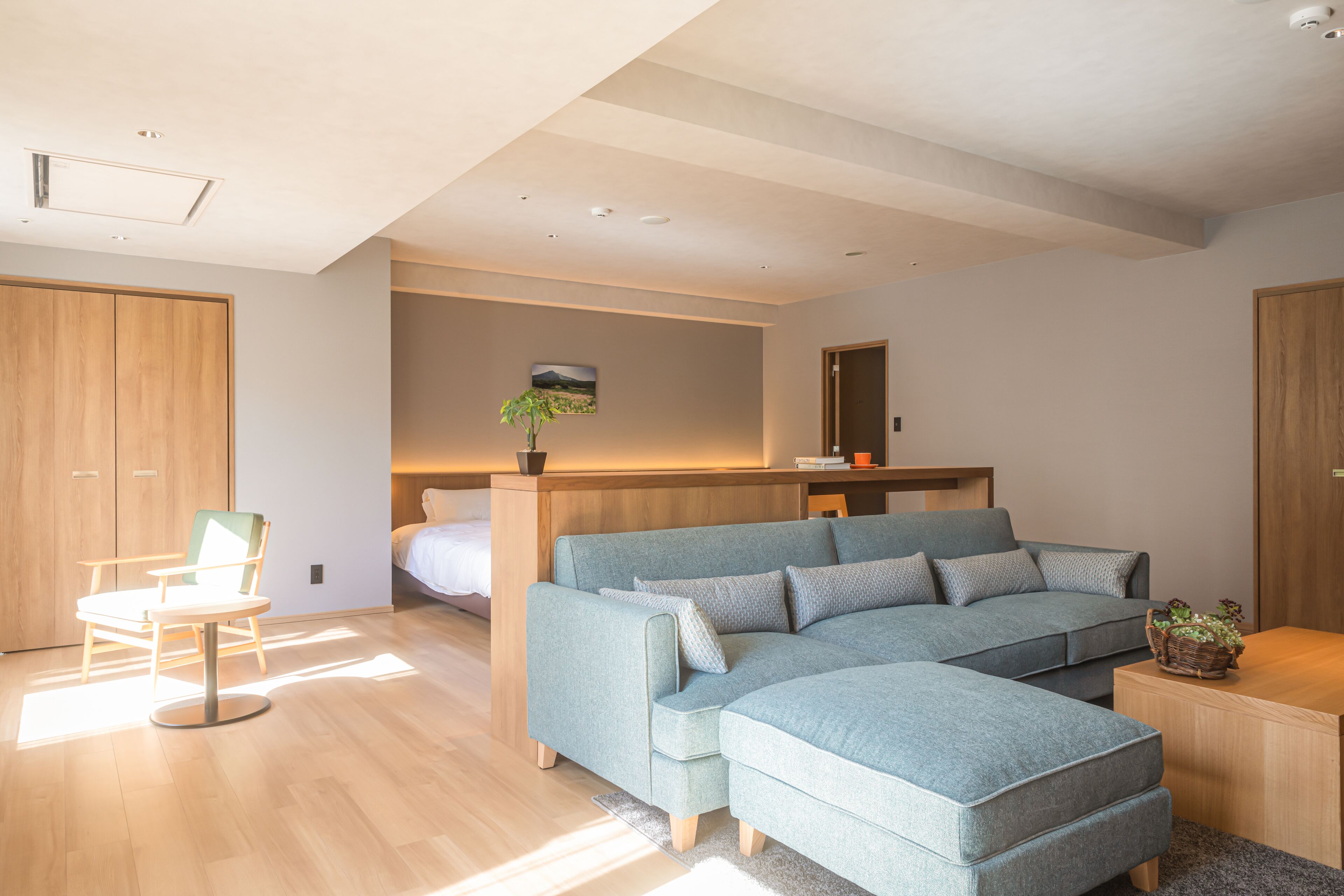 A modern bedroom with a light blue sectional sofa, a wooden wardrobe, a bedside table with a plant, and a large mirror above the bed.