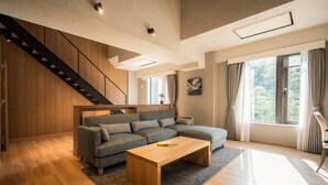 Un salon moderne avec un escalier en bois, un canapé d’angle, une table basse en bois et de grandes fenêtres avec des rideaux.