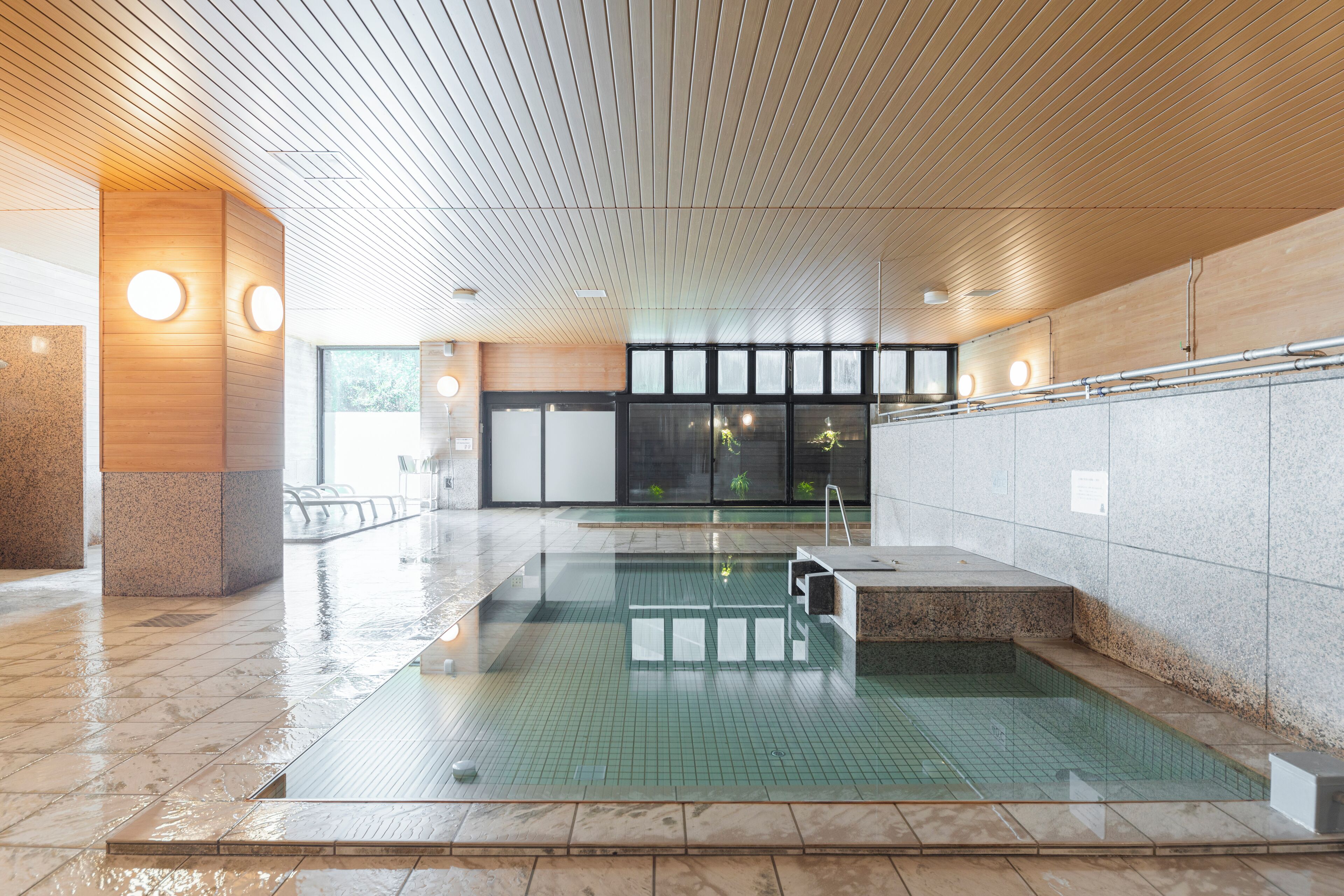 A swimming pool with a tiled floor and a wooden ceiling, surrounded by walls with a stone finish and a glass door leading outside.