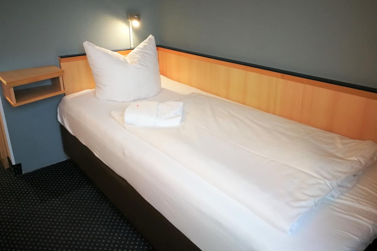 A neatly made bed with white linens and pillows, a wooden headboard, and a bedside table with a lamp.