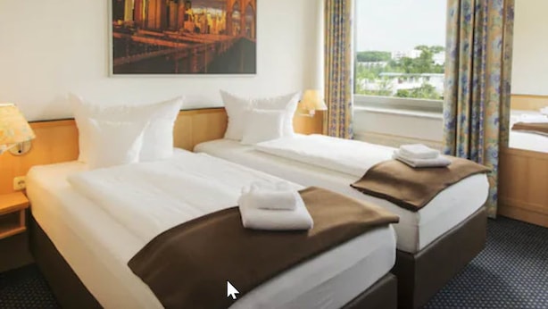 A hotel room with two beds, a cityscape painting, a window with curtains, and a view of greenery. - Hotel NearBy (Laatzen)