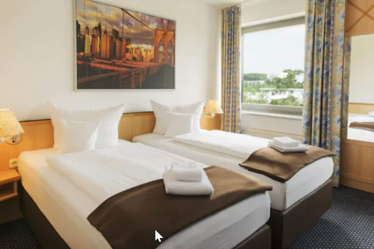 A hotel room with two beds, a cityscape painting, a window with curtains, and a view of greenery.