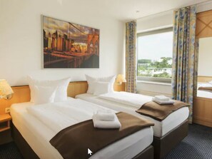 A hotel room with two beds, a cityscape painting, a window with curtains, and a view of greenery. - Hotel NearBy (Laatzen)