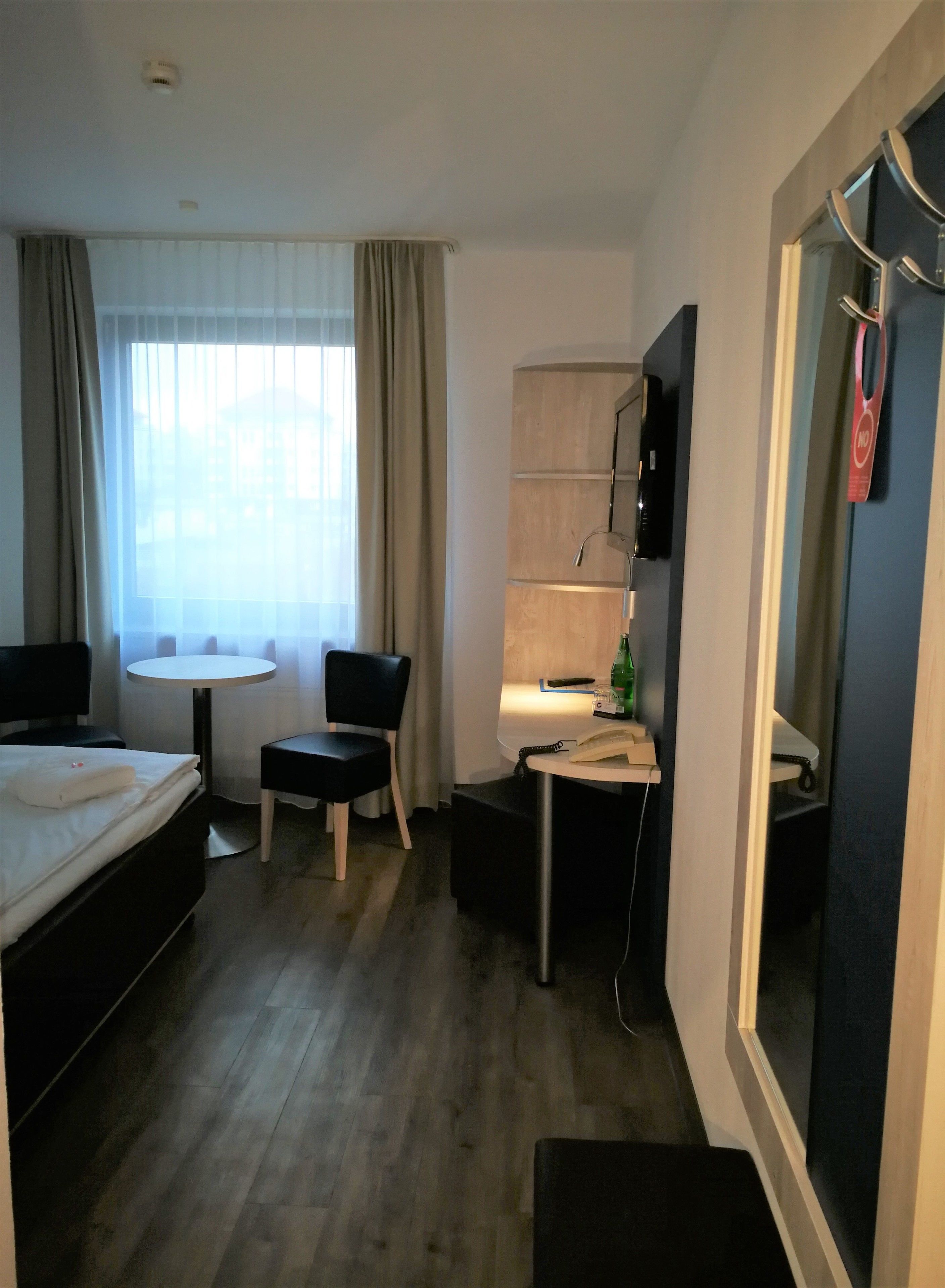 Quarto de hotel com cama, escrivaninha, cadeira, mesa pequena, televisão e janela com cortinas.