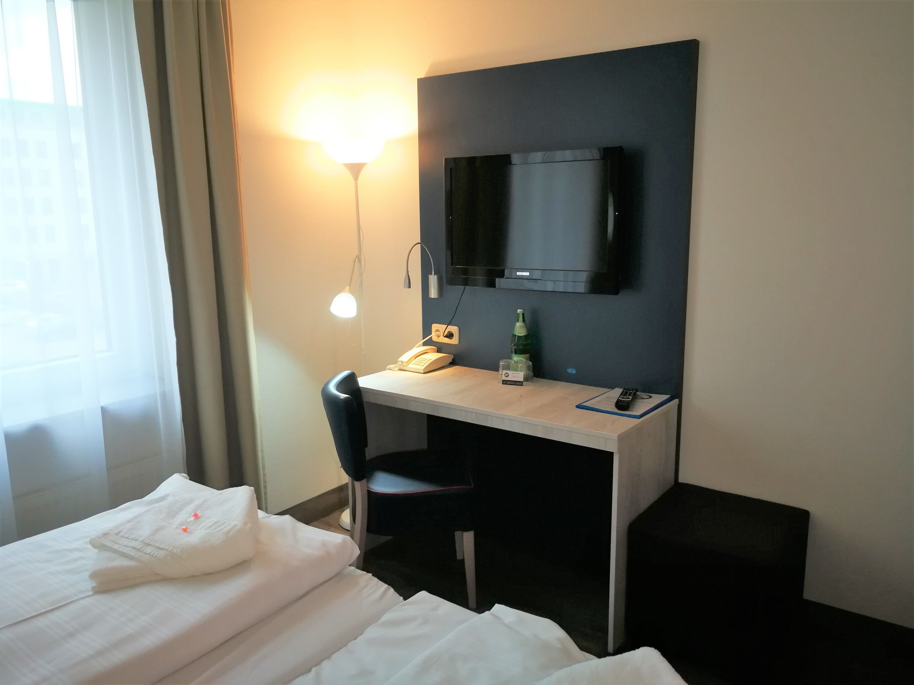 Una habitación de hotel con cama, escritorio con silla, teléfono, una botella y televisor montado en la pared.