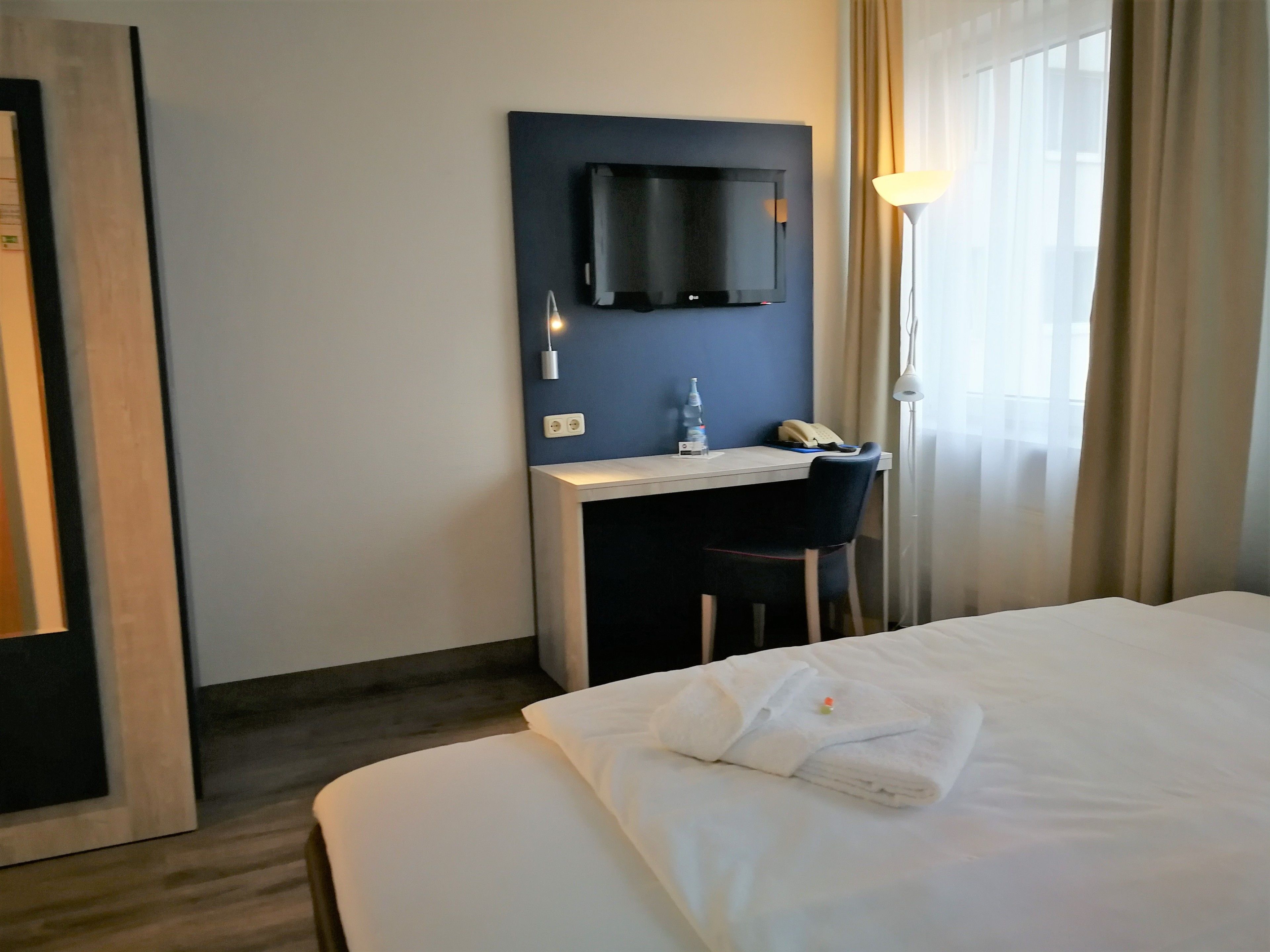 Ein Hotelzimmer mit einem Bett, einem Schreibtisch mit Stuhl, einem flachbildfernseher an der Wand, einer Lampe und einem Fenster mit Vorhängen.