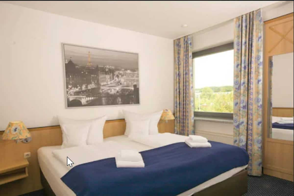 A hotel room with a large bed, a cityscape painting, a window with curtains, and a nightstand with a lamp.
