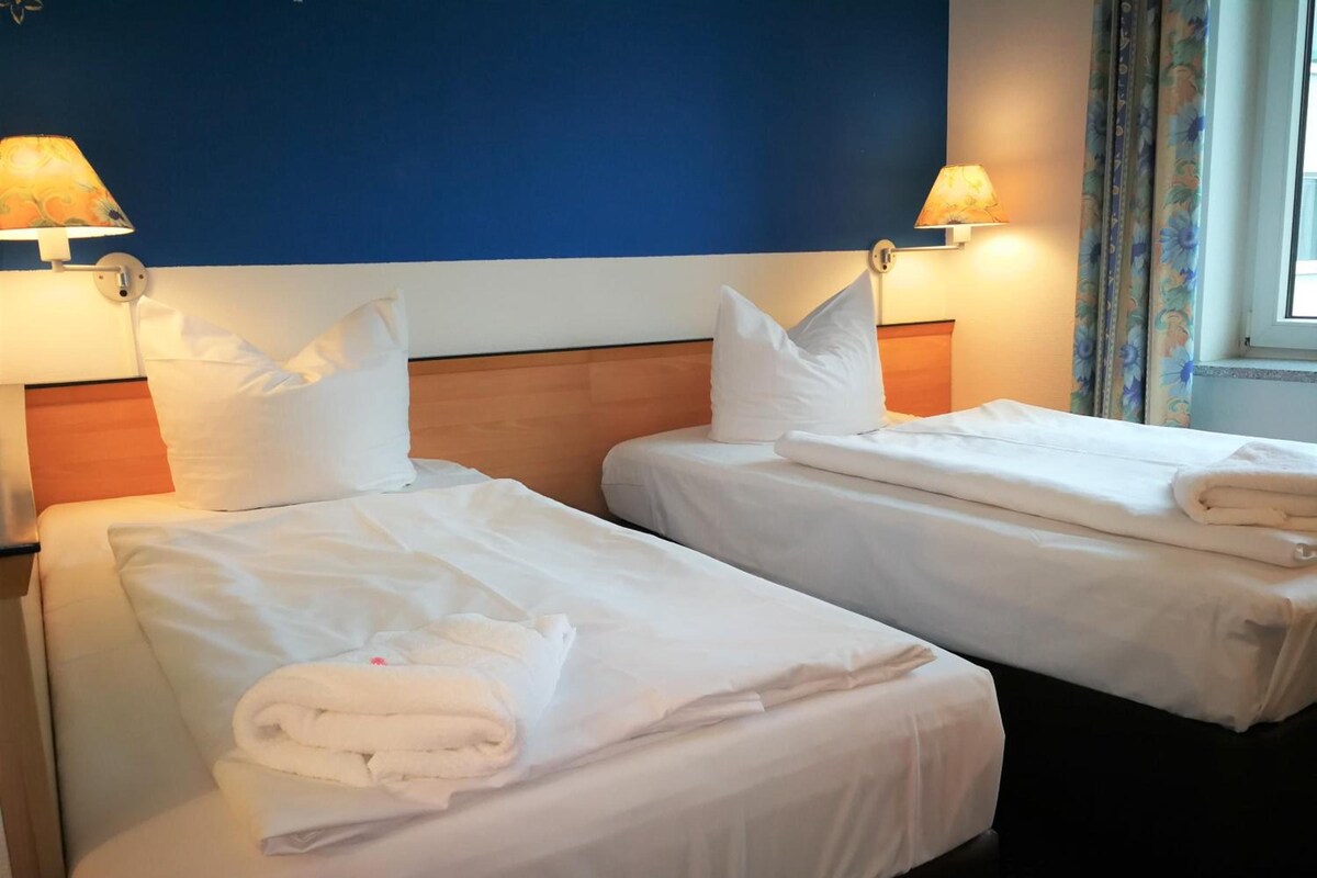 A hotel room with two beds, each with white linens and pillows, a wooden headboard, and two wall-mounted lamps.