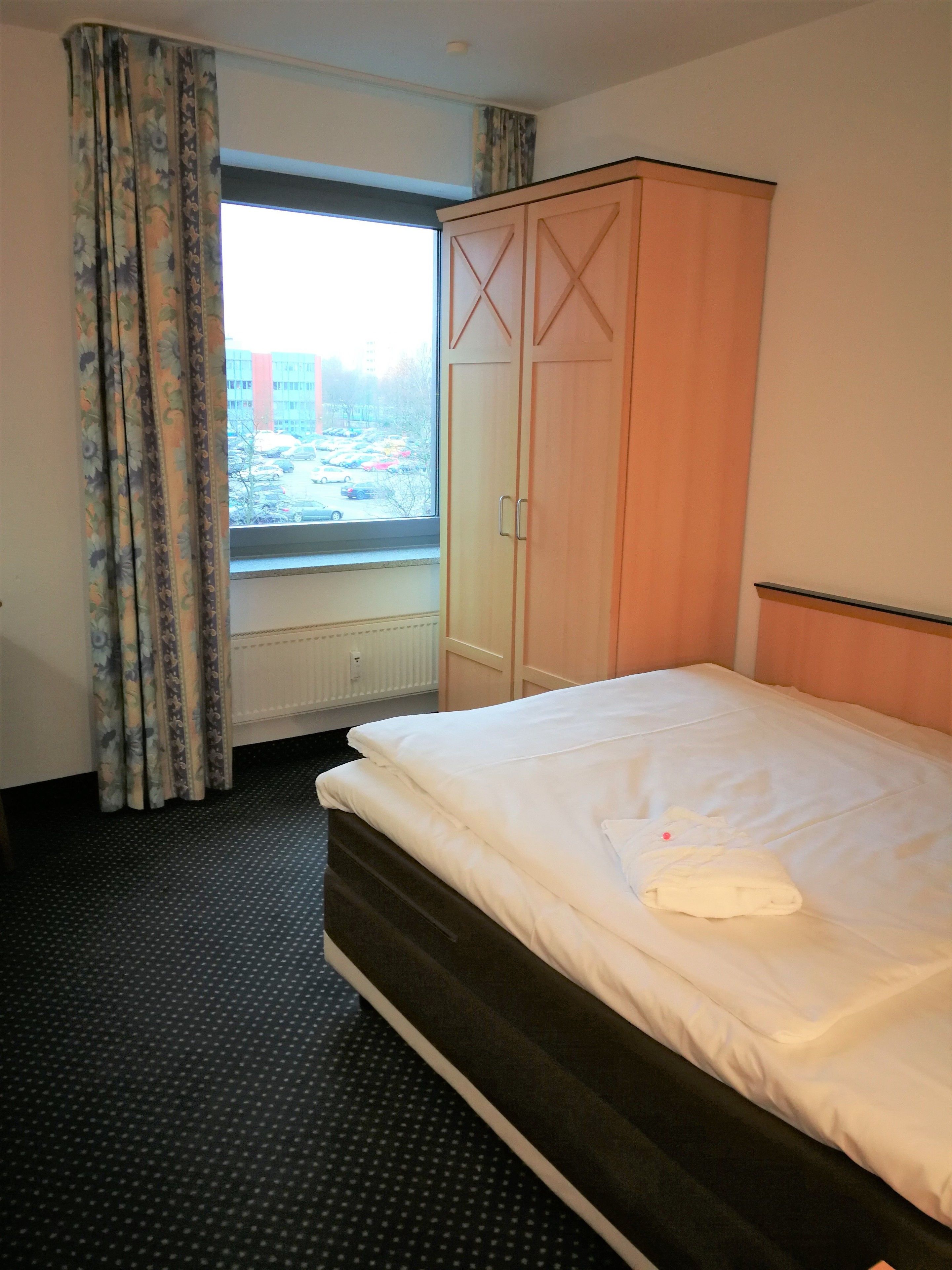 Habitación de hotel con cama, armario, ventana con cortinas y vistas al exterior.