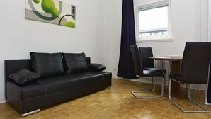 A modern living room with a black leather sofa, a dining table with chairs, a window with curtains, and a painting on the wall.