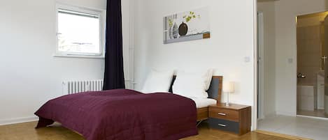 Un dormitorio con piso de madera, una cama con cubrecama morado, una mesita de noche con lámpara, una ventana con cortina y un cuadro en la pared.