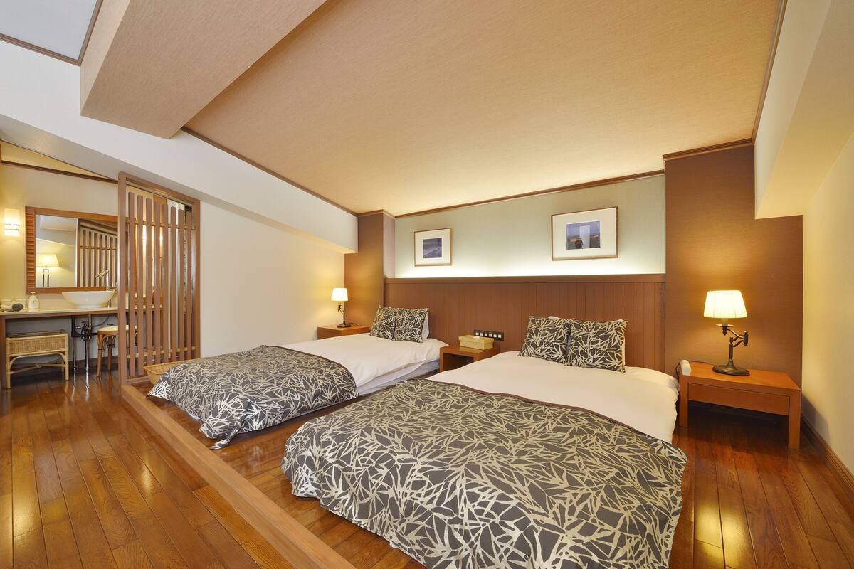 Habitación de hotel con dos camas, suelo de madera, mesitas de noche con lámparas, un escritorio con silla y un espejo.