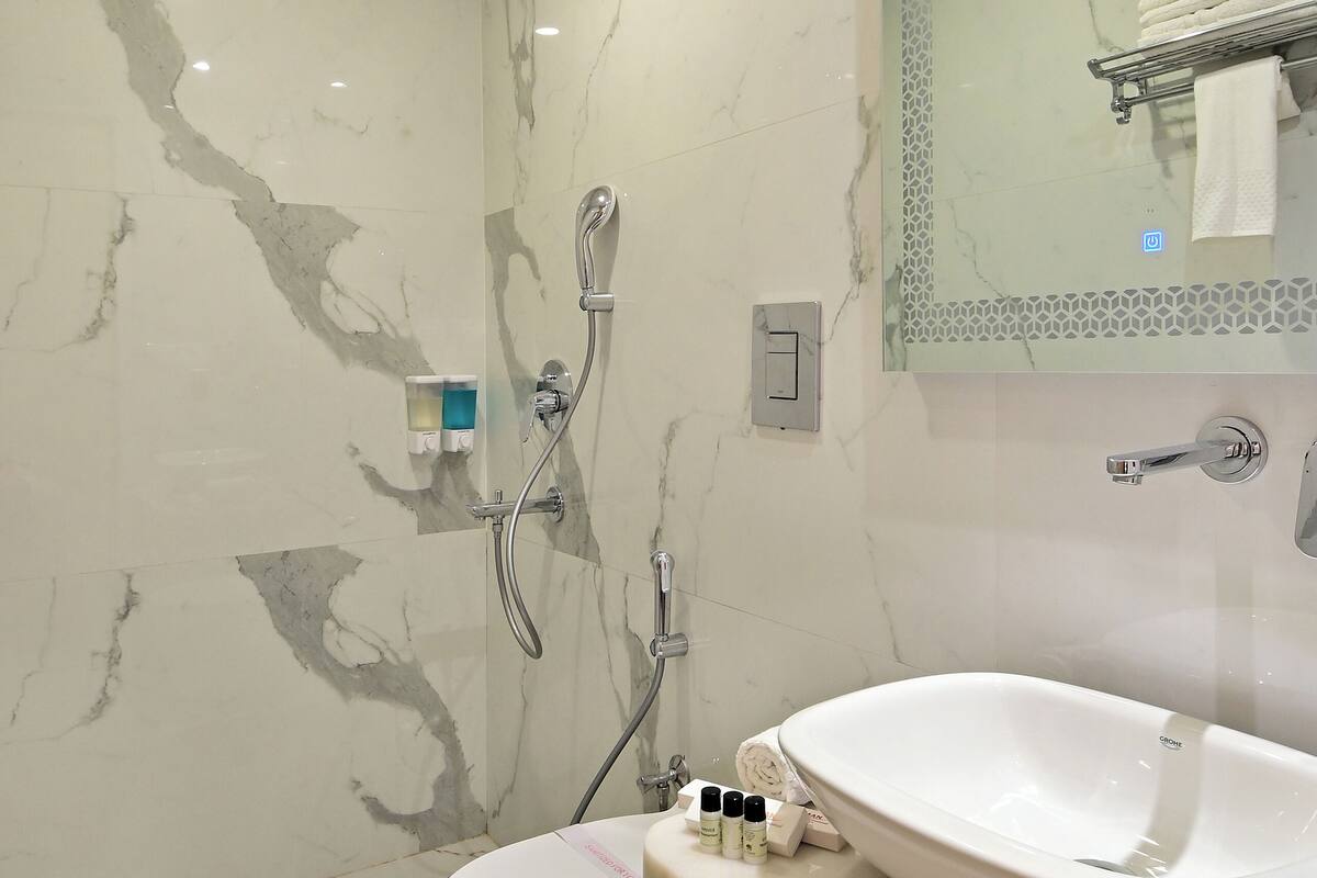 Un baño con lavamanos, inodoro y zona de ducha, paredes de mármol y un espejo con marco decorativo.