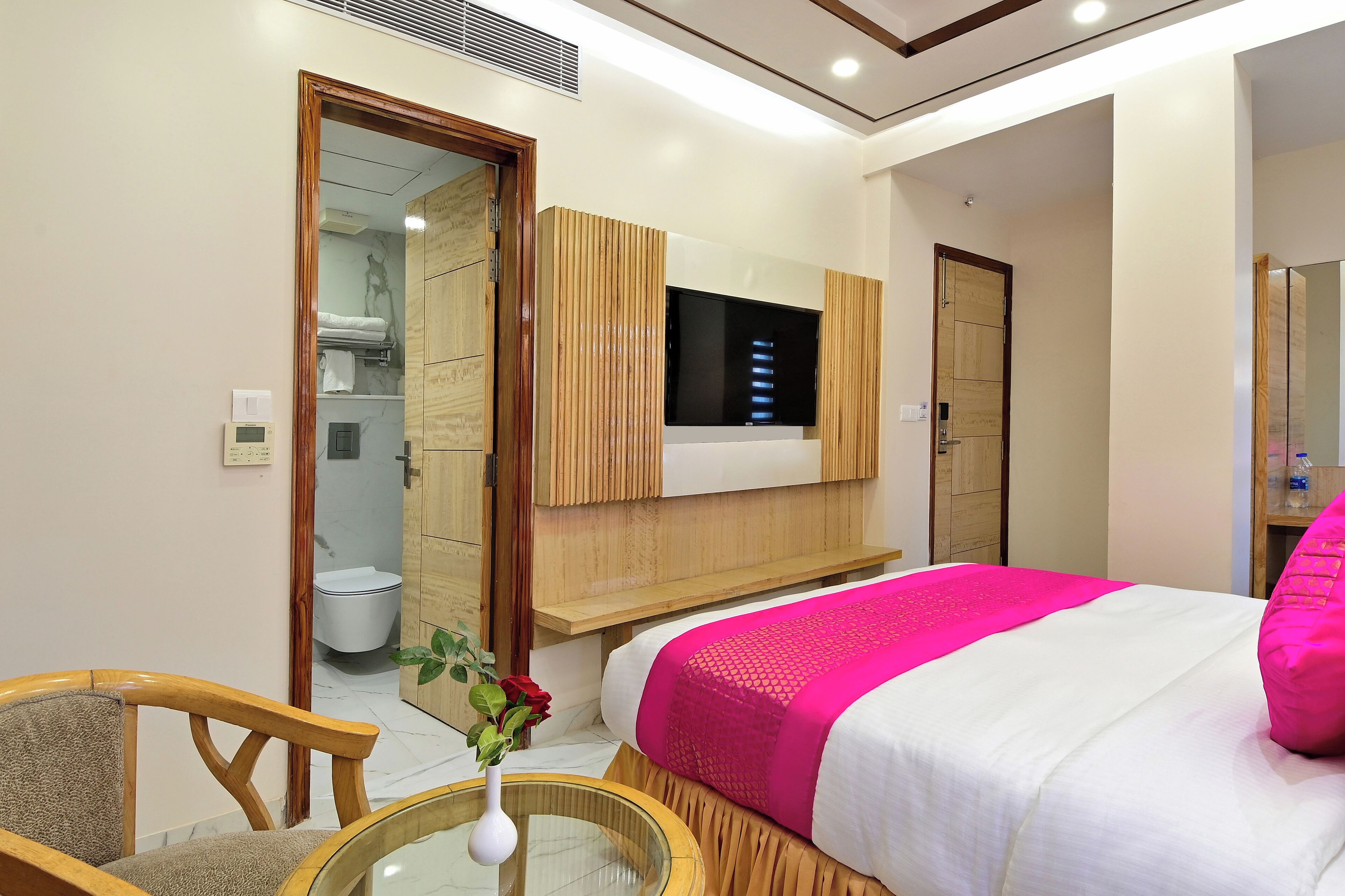A modern hotel room with a large bed, a TV mounted on the wall, a wooden vanity, and a bathroom visible through an open door.