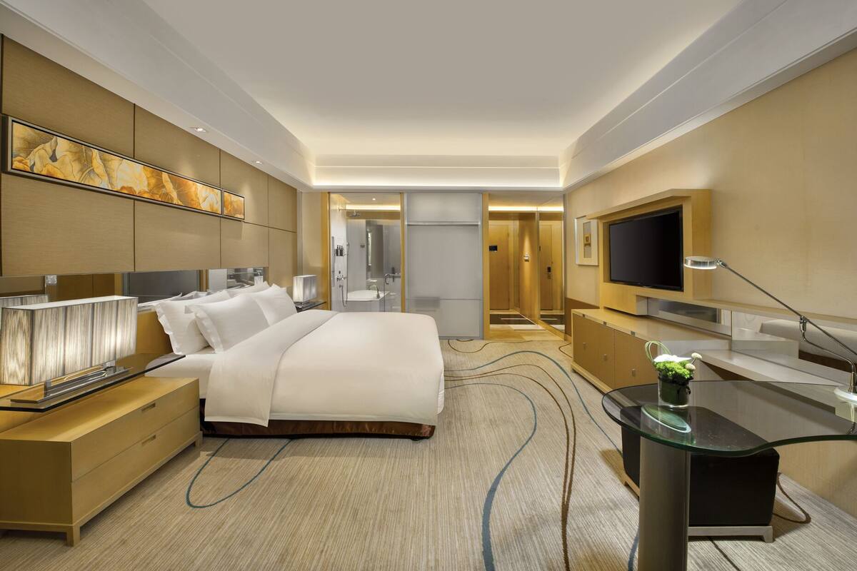 Una habitación de hotel moderna con una cama grande, un escritorio, un televisor y un baño visible a través de un panel de vidrio.