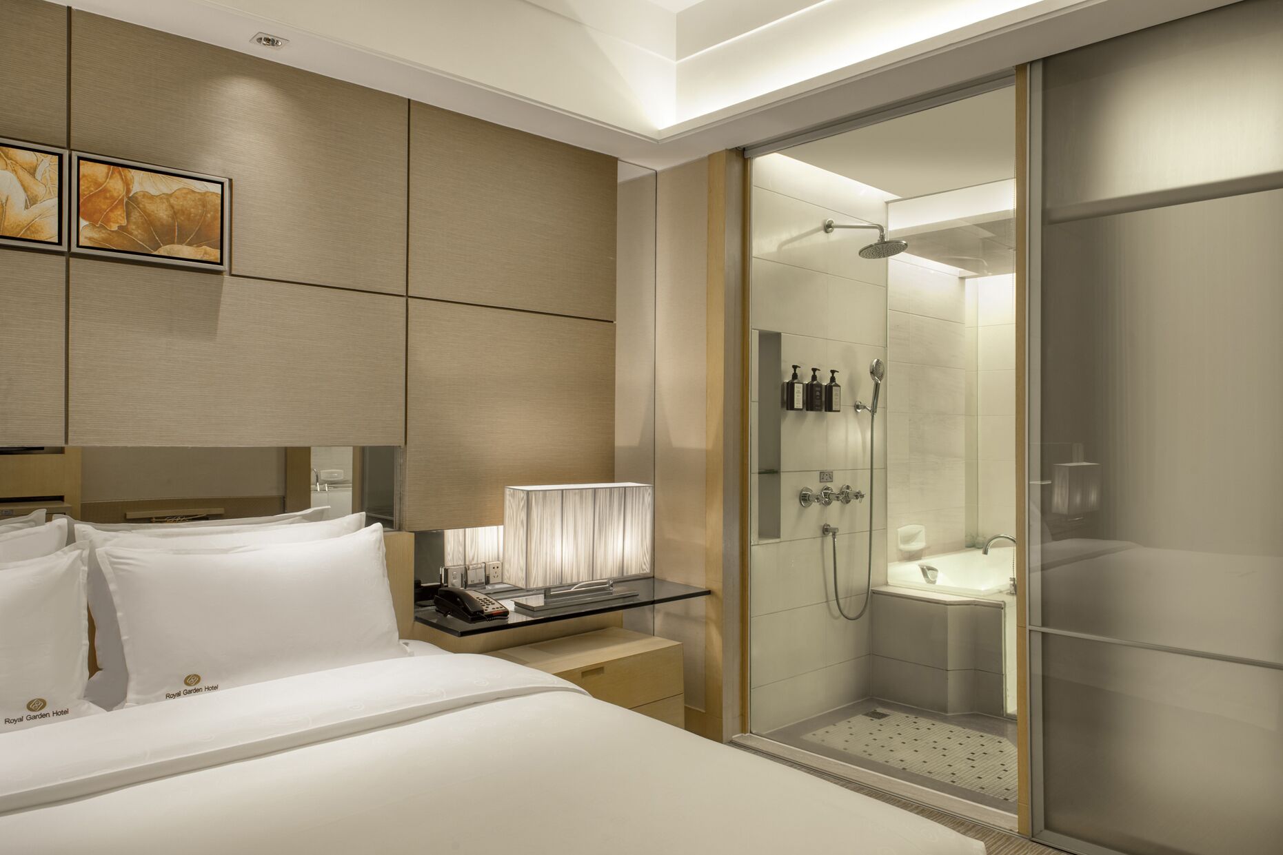 A hotel room with a bed, bedside table, lamp, and a glass-enclosed shower area.