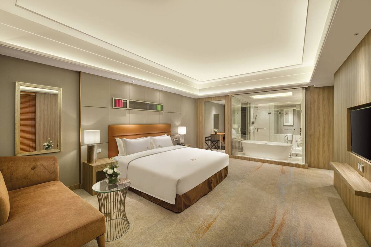Habitación de hotel moderna con una cama grande, una zona de estar, un baño con paredes de vidrio y un televisor.