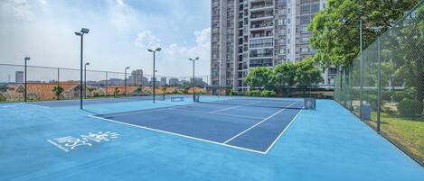 Una cancha de tenis exterior con superficie azul, rodeada por una cerca de malla metálica y contigua a un edificio residencial alto.