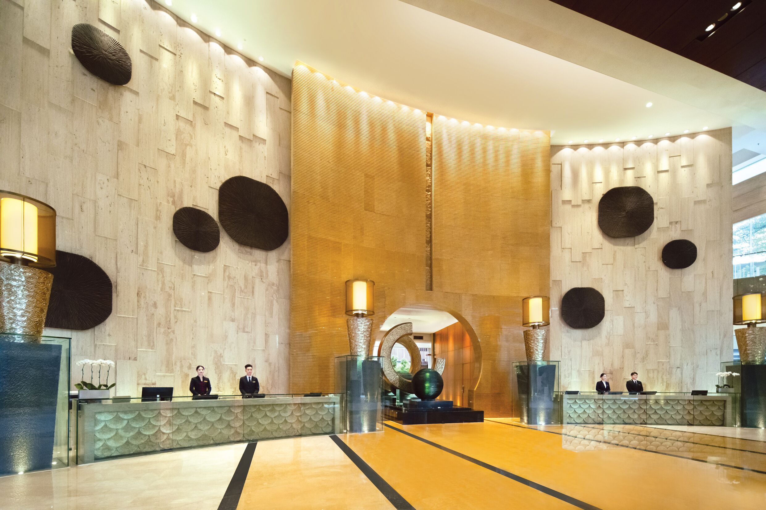 Un espace d’accueil au design moderne, comprenant un comptoir de réception, deux réceptionnistes, des panneaux muraux décoratifs et un grand élément décoratif mural illuminé.