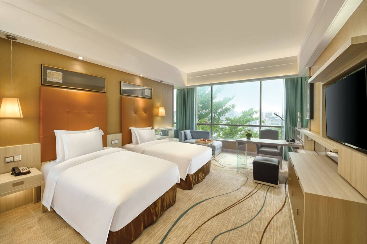 Habitación de hotel con dos camas, un escritorio, televisión y balcón con vista a la ciudad.