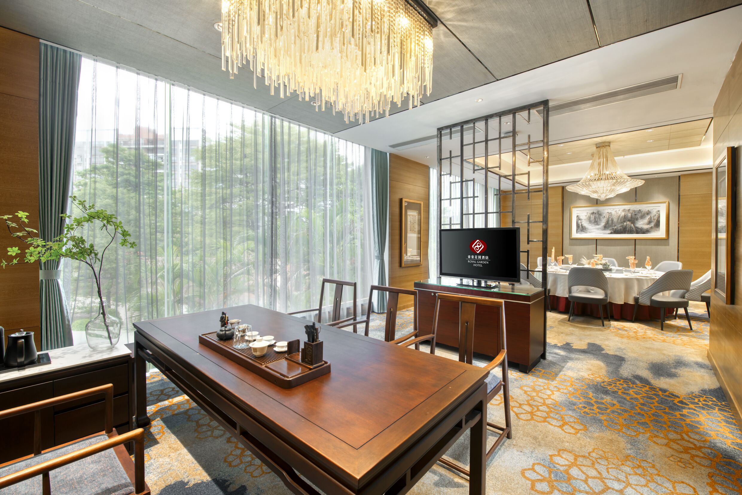 A hotel room with a wooden dining table, chairs, a TV, and a large window with curtains.