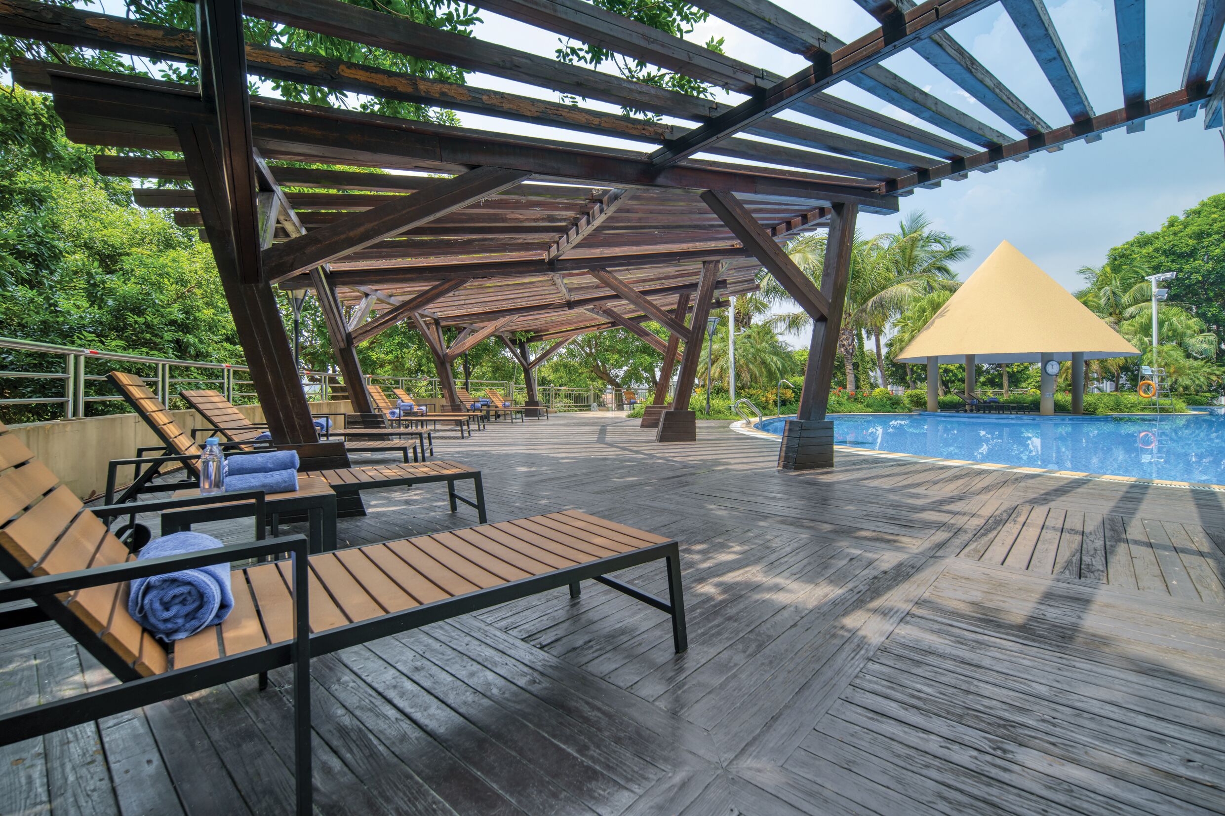 Terraza de madera con sillones de descanso y un área cubierta con techo amarillo, con vista a una piscina.