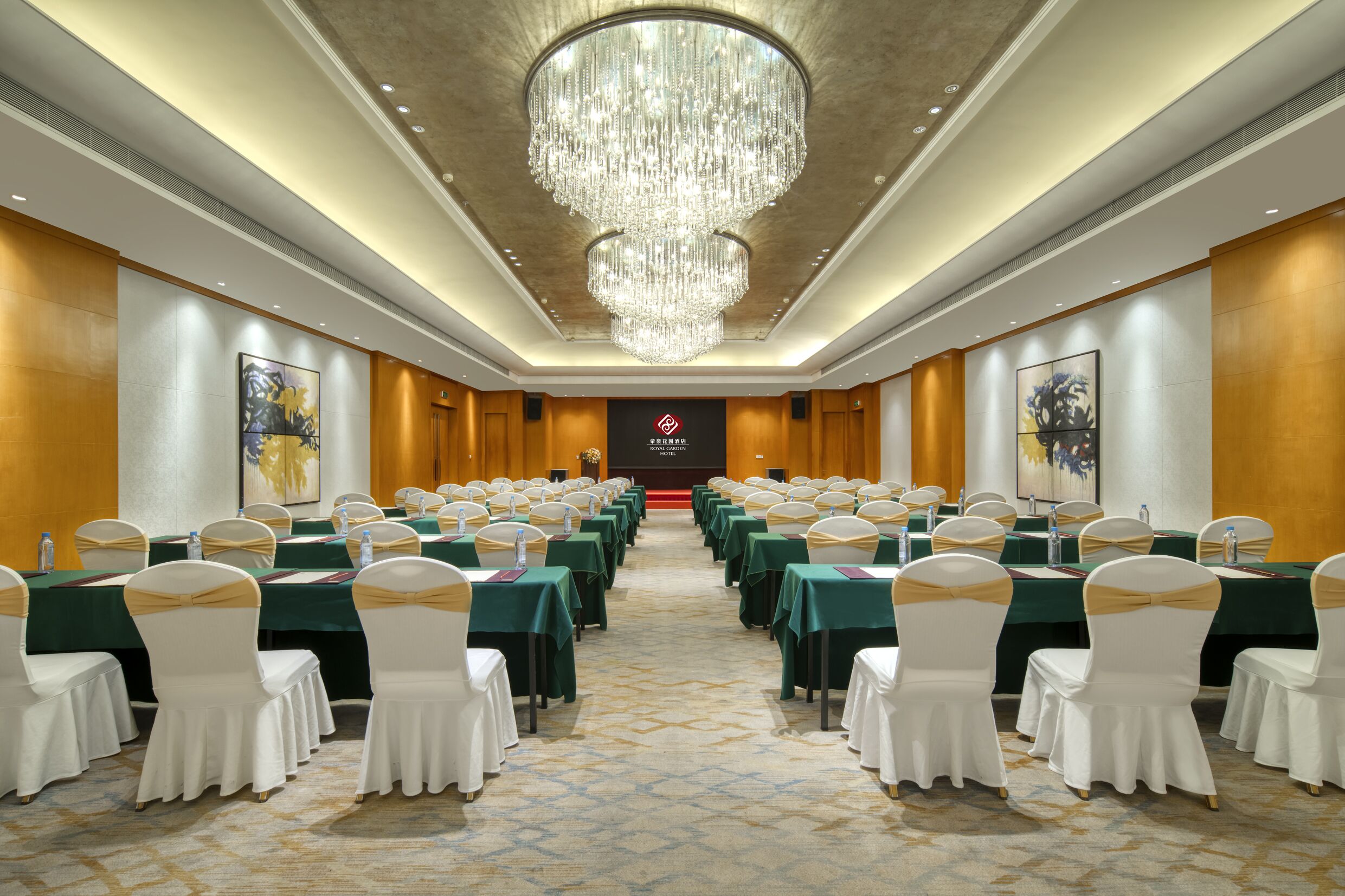 A conference room with rows of tables and chairs, a chandelier, and a stage with a logo.